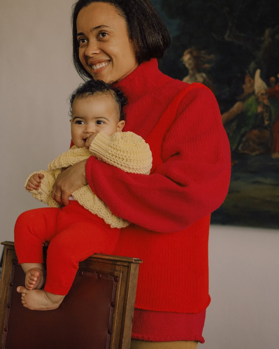 A smiling mother in a red sweater holds her baby in a yellow sweater while sitting on a chair in an indoor environment with a painting.