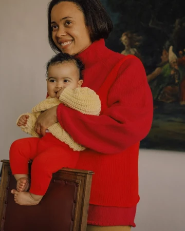 A smiling mother in a red sweater holds her baby in a yellow sweater while sitting on a chair in an indoor environment with a painting.