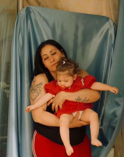 A woman with dark hair and a tattoo holds a toddler wearing a red dress in front of a blue curtain, indoors, in a medium shot.