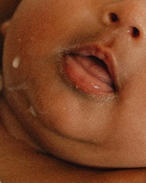 Close-up of baby's face with open mouth, showing tongue and lips, alongside dribbles of liquid on the skin and small nostrils.