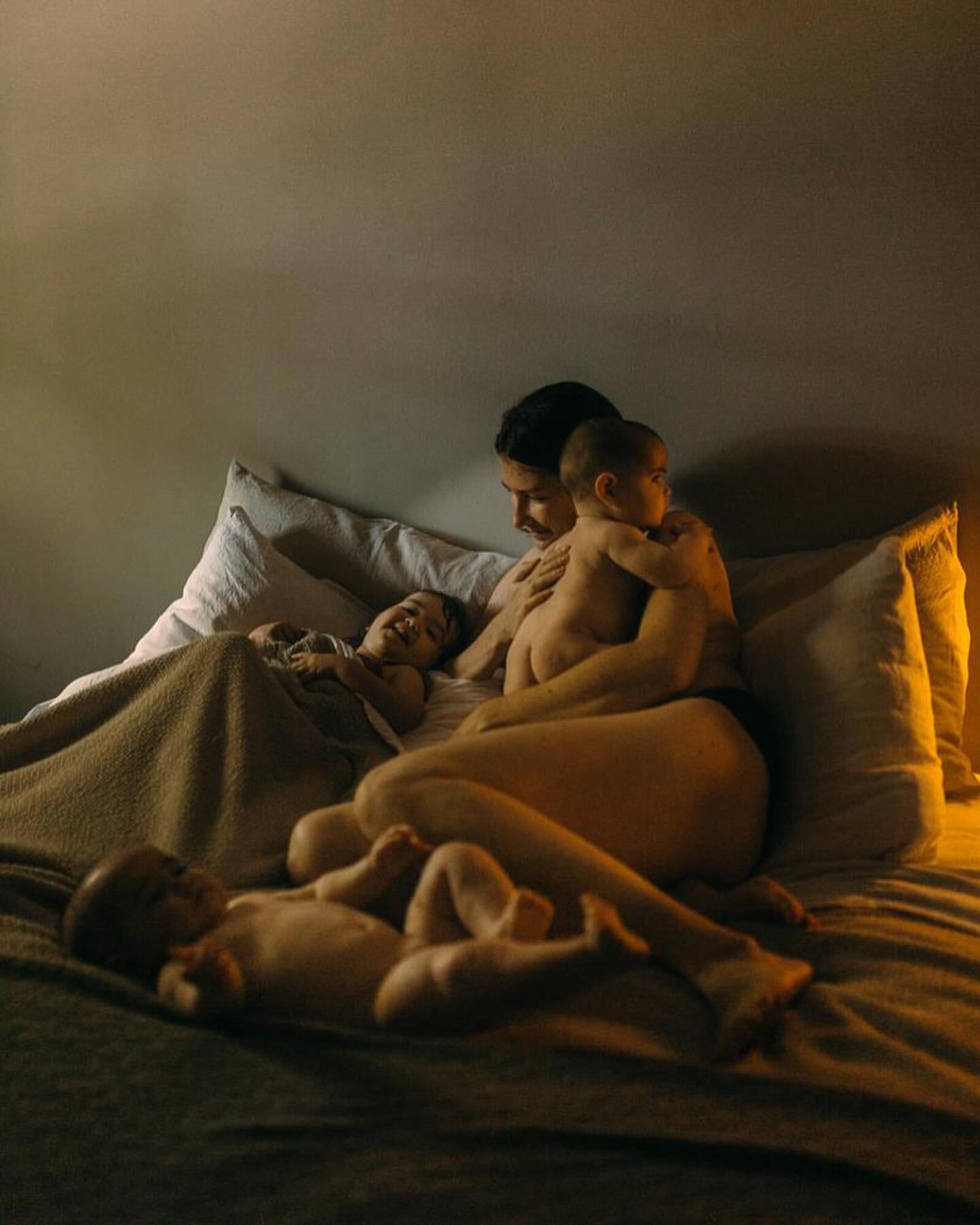 Indoor shot of a mother sitting on a bed with three children. Warm lighting emphasizes the intimate, familial scene in the bedroom.