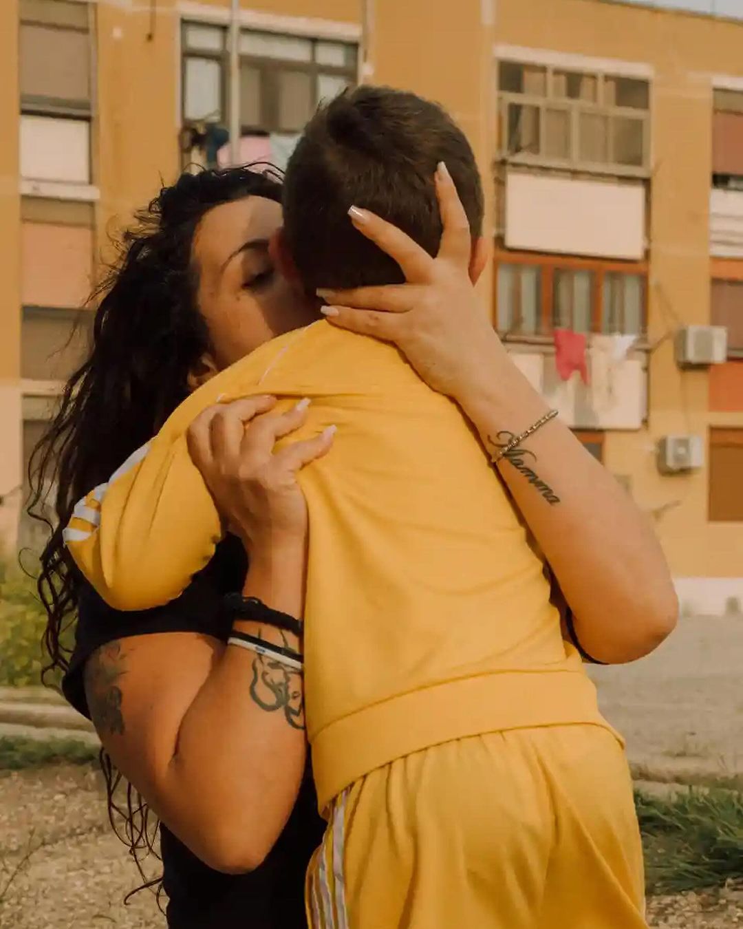 Adult female hugs child outdoors. Woman with dark hair and tattoos embraces the child, both wearing matching yellow tracksuits.