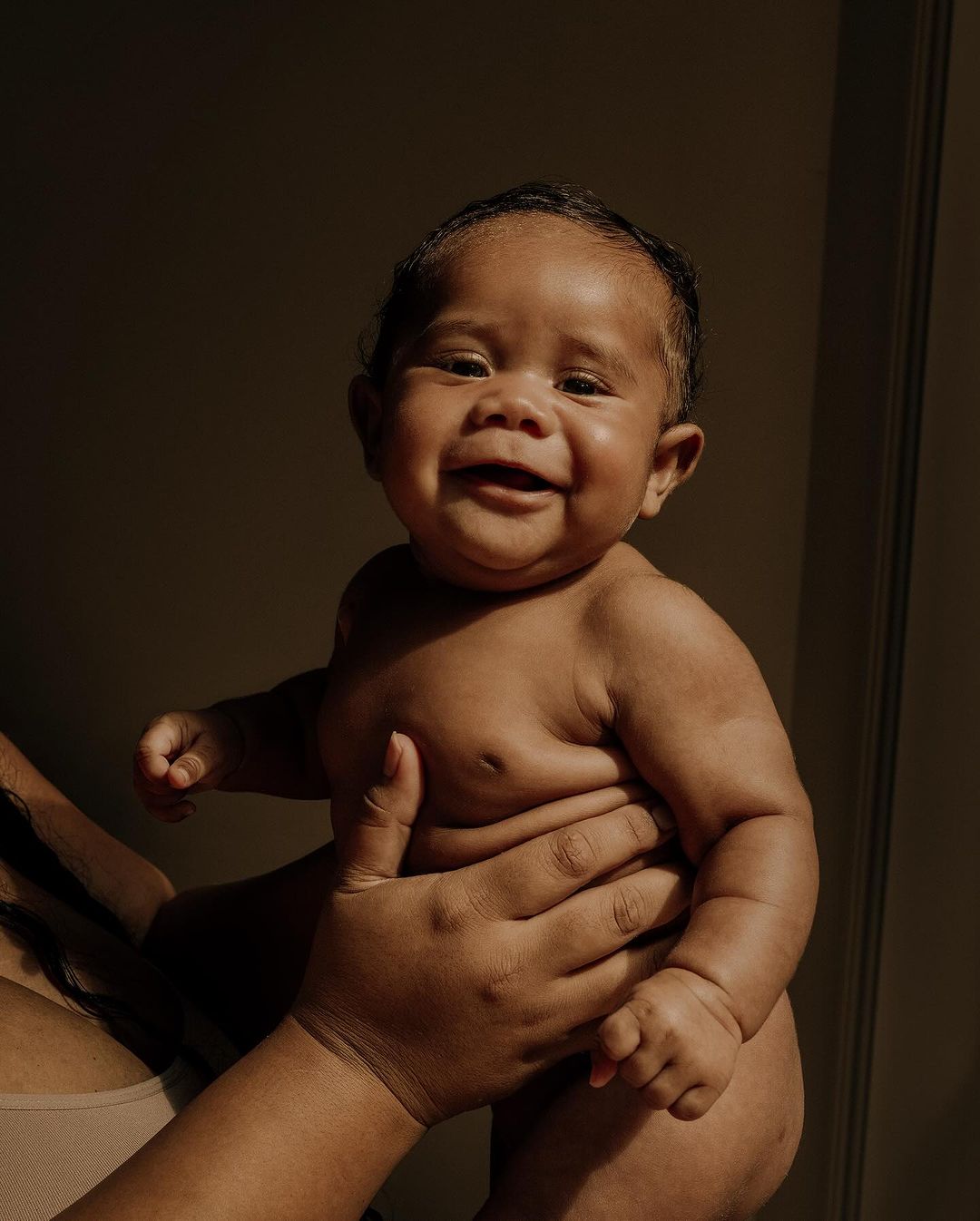 Close-up of a happy infant being held up. The baby has dark skin and dark hair. An adult holds the baby with their hands.