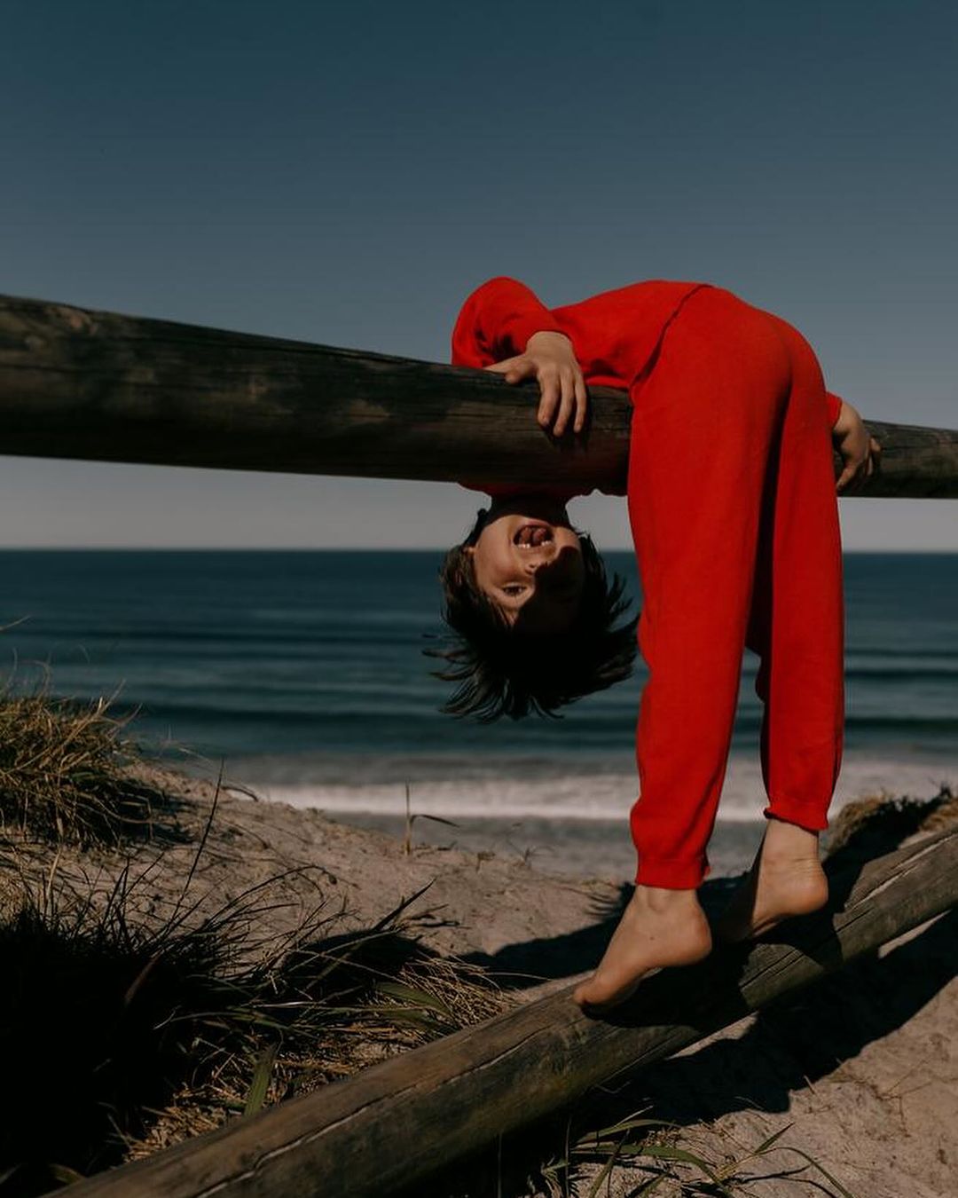 Child in red outfit playfully hangs upside down on a wooden fence near the beach with blue sky and ocean backdrop in a candid shot.