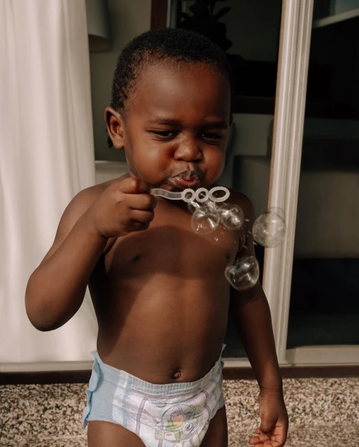 Toddler with dark hair wearing a diaper blows bubbles indoors, captured in a medium shot showcasing their playful activity.