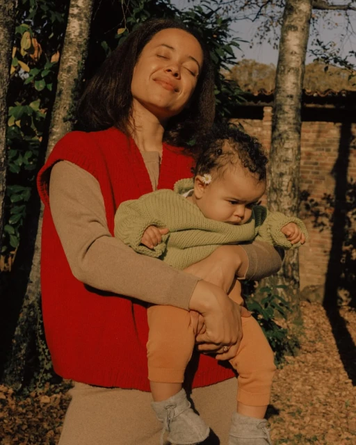 A woman holds a baby outdoors near trees and a brick wall. The mother wears a red vest and beige sweater, the baby wears green and orange.