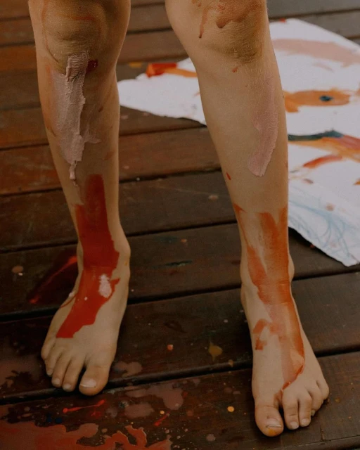 Child's legs and feet covered in paint standing on a wooden deck, next to a painting also covered in red, orange, and white paint.