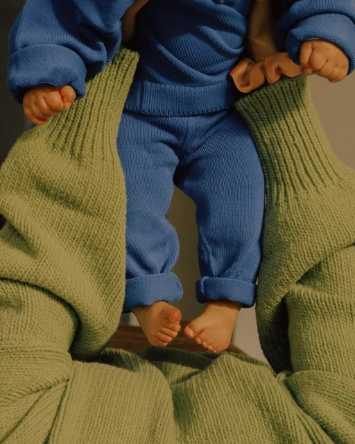 Closeup of a baby held up by someone in a green knitted sweater; the baby is wearing a blue knit suit, indoor setting, natural lighting.