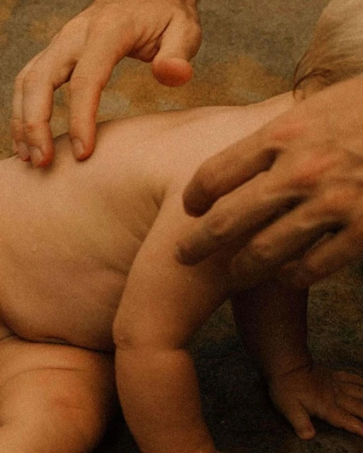 Close-up of a baby's back and buttocks being touched and examined by the hands of an adult with a warm, neutral-toned background.