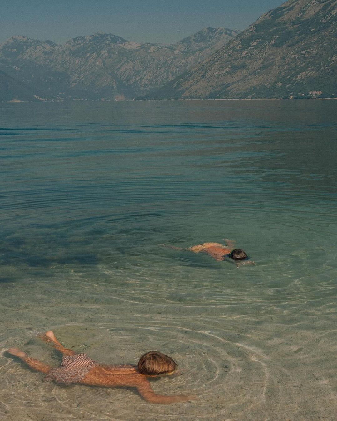 Two children swimming in shallow clear water with distant mountains rising across a calm lake under daylight.
