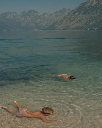 Two children swimming in shallow clear water with distant mountains rising across a calm lake under daylight.