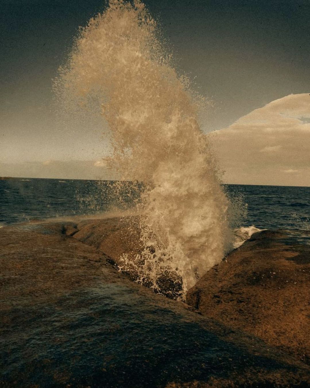 Ocean wave crashes against a rocky outcropping, creating a dynamic display of water and power under an overcast sky, landscape orientation.
