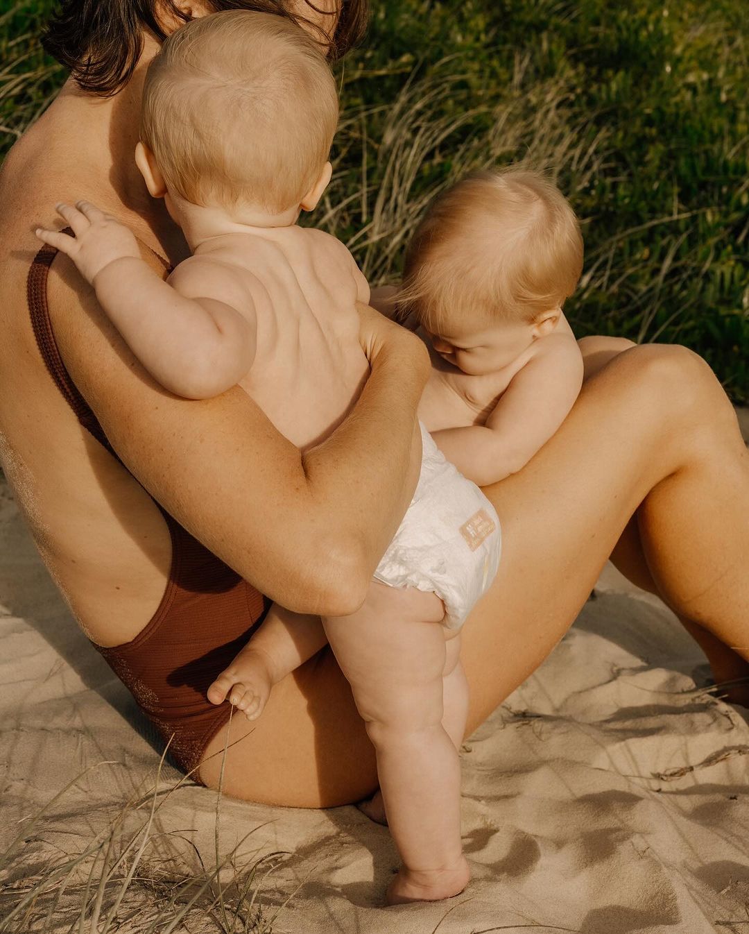 A mother in a brown swimsuit holds two babies in diapers against a backdrop of sand and grassy foliage, shot in warm, natural light.