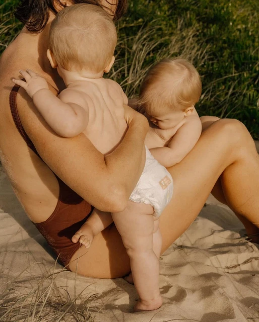 A mother in a brown swimsuit holds two babies in diapers against a backdrop of sand and grassy foliage, shot in warm, natural light.