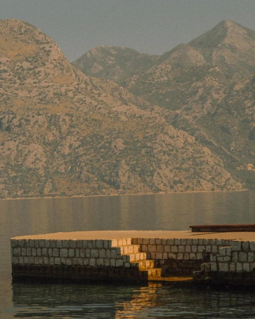 Coastal scene featuring a stone pier with stairs leading into the water, set against a backdrop of mountains and serene water.