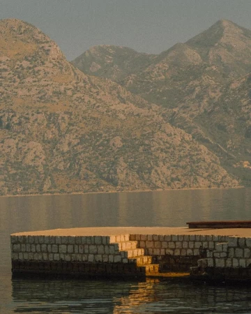 Coastal scene featuring a stone pier with stairs leading into the water, set against a backdrop of mountains and serene water.