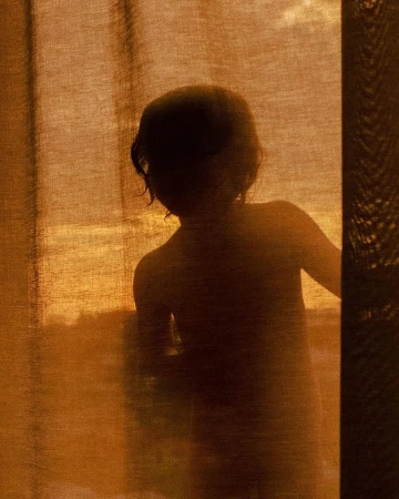 Silhouette of a child behind a sheer warm-toned curtain with soft golden light filtering through the fabric.