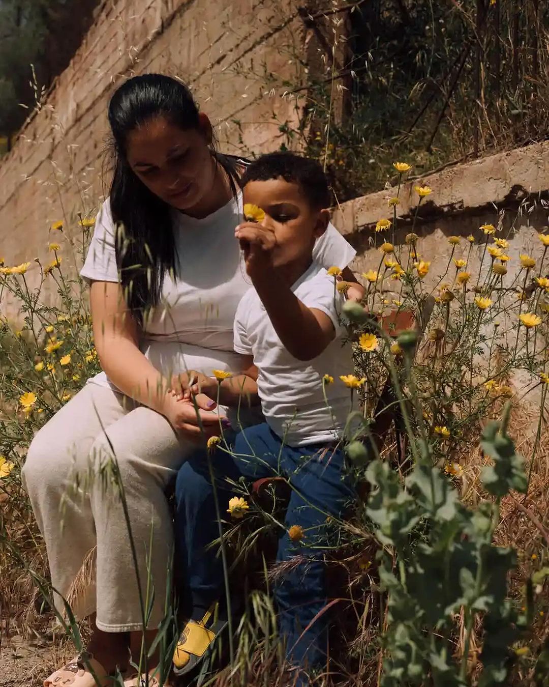 A mother and child are sitting together among yellow flowers in a field. The child is holding a flower and looking at it closely.