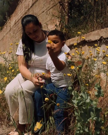A mother and child are sitting together among yellow flowers in a field. The child is holding a flower and looking at it closely.
