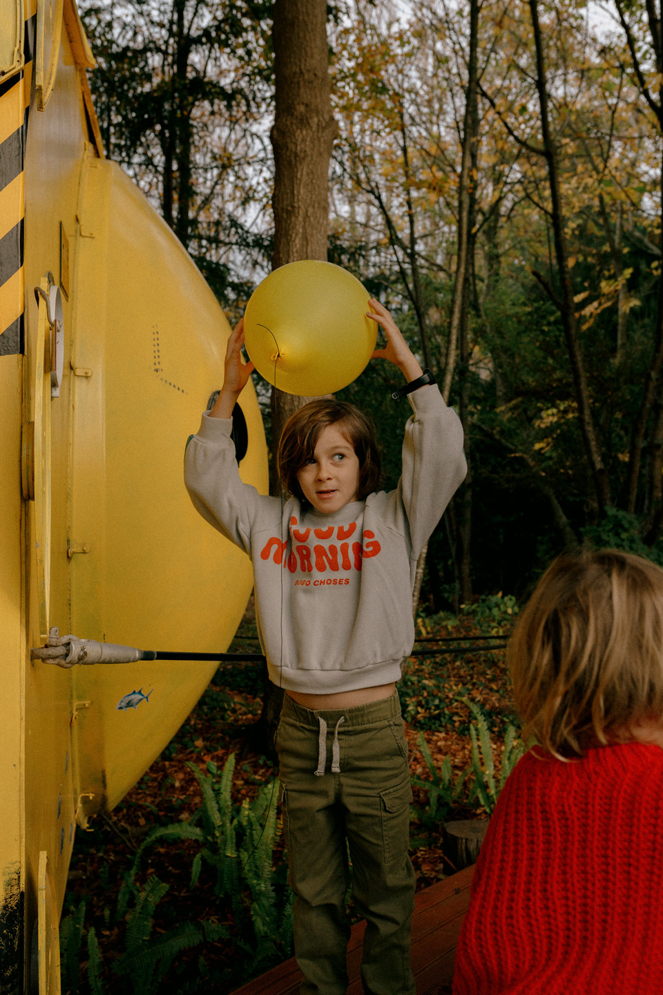 A young child stands outdoors in front of yellow structure, holding a yellow balloon up over their head, with trees in the background.