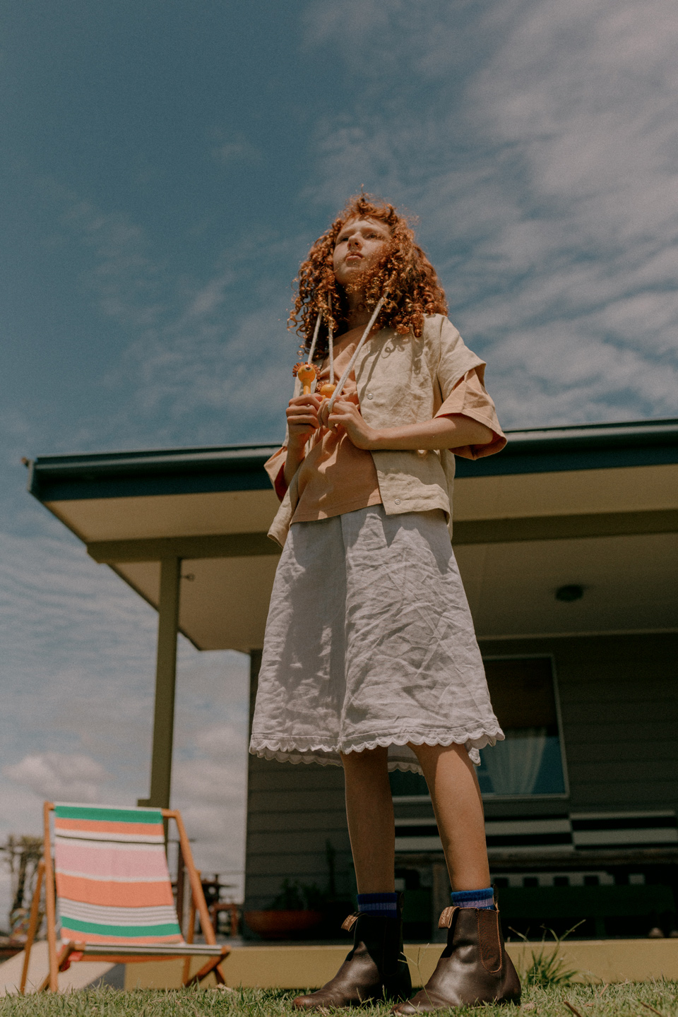 Young woman with curly red hair stands outside a house wearing a light jacket and skirt, holding a pair of orange-handled objects.