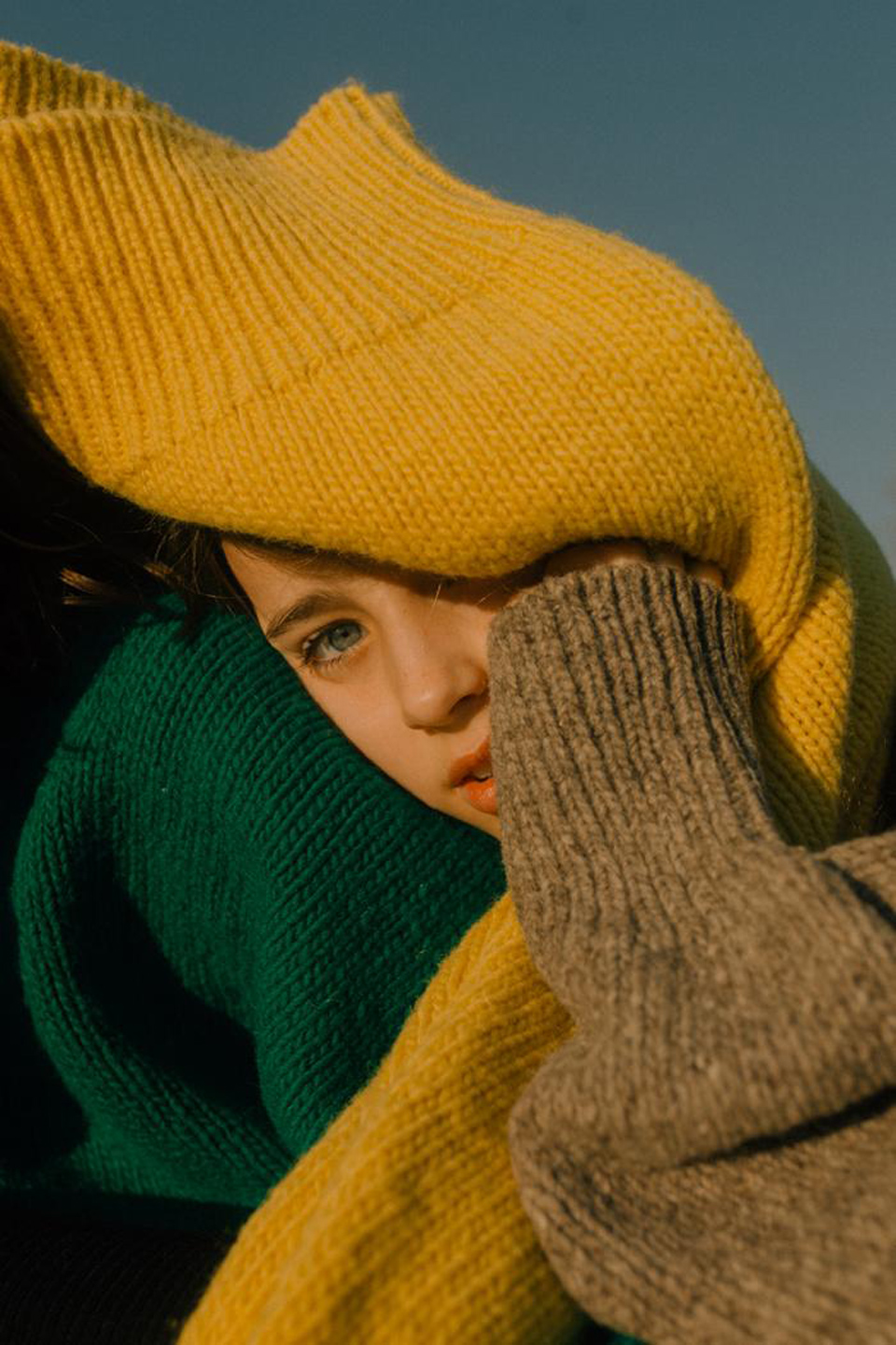 Close-up of a child's face framed by colorful knit sweaters in yellow, green, and grey, against a clear sky background.