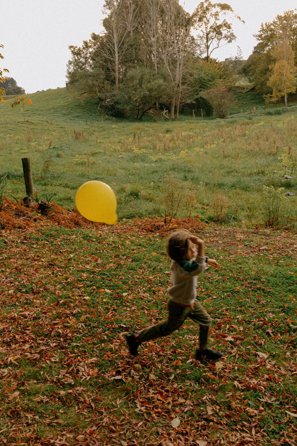 A child runs across a lawn covered in fallen leaves, chasing a yellow balloon, with a grassy hill and trees in the background.
