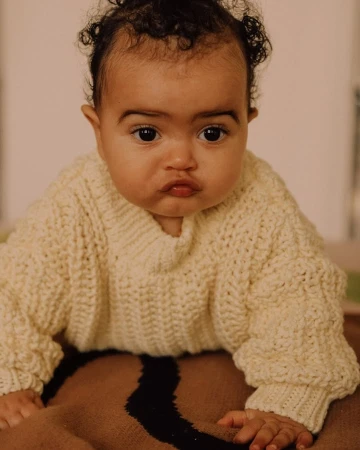 A cute baby with brown eyes and dark curly hair, wearing a cream-colored knitted sweater, is lying on a brown blanket with a black stripe.