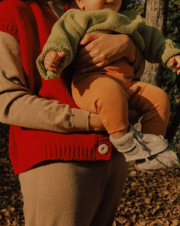 A baby wearing a green sweater is being held by an adult in a red cardigan in an outdoor setting with trees and leaves in the background.