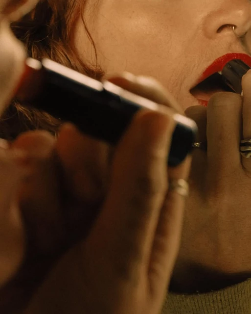 Close-up shot of fair-skinned adult female applying bright red lipstick with a dark applicator tube. The background is blurred.