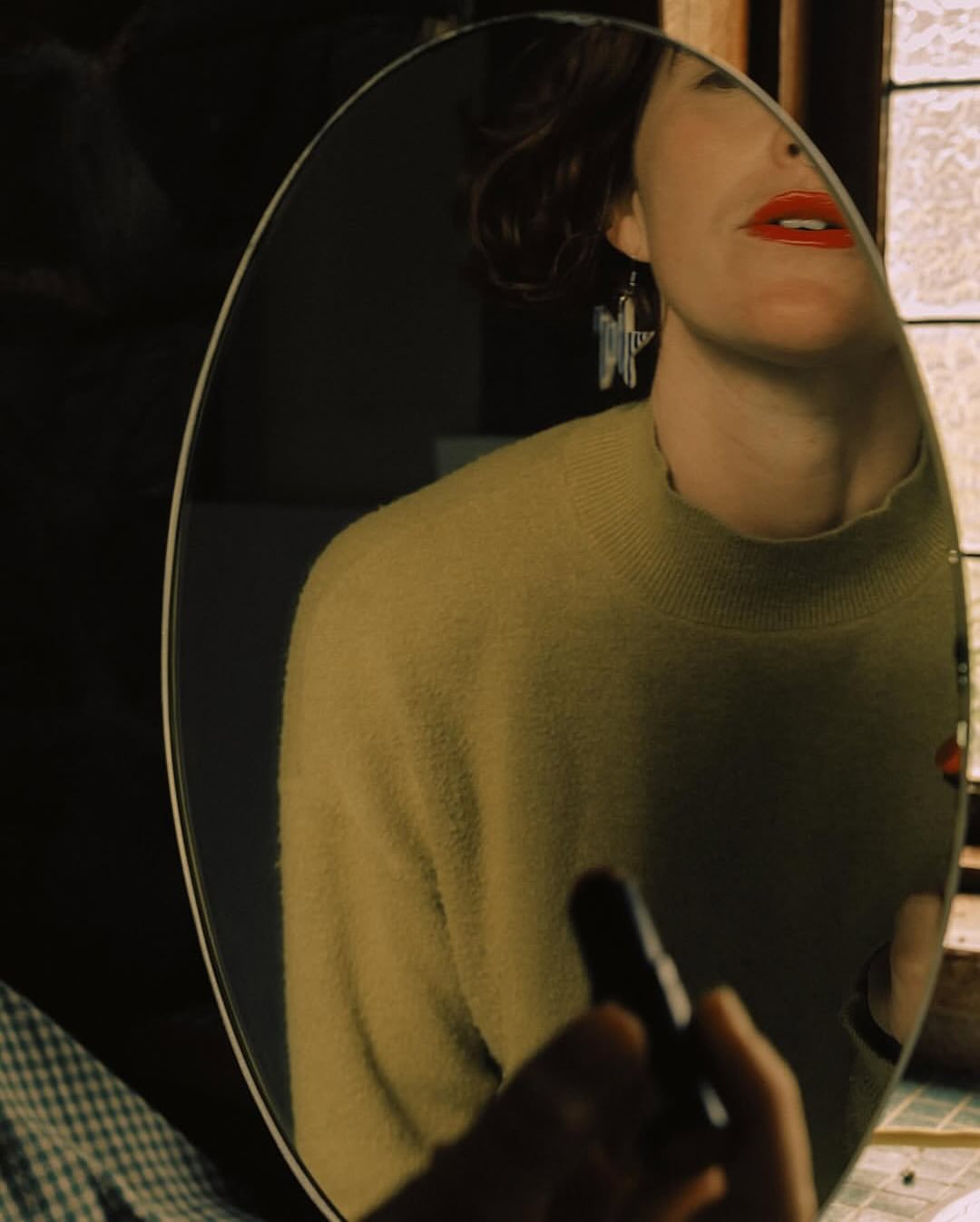 Reflection in an oval mirror shows a woman applying red lipstick, wearing a yellow sweater and star earrings in a softly lit room.