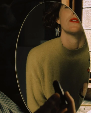 Reflection in an oval mirror shows a woman applying red lipstick, wearing a yellow sweater and star earrings in a softly lit room.