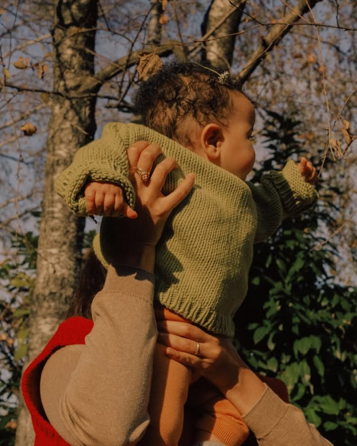 A baby in a green sweater is being held up outdoors by a woman in a tan sweater, with trees and a blue sky in the background.
