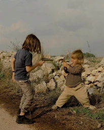 Two children playing outdoors with wooden sticks against a rocky ground and overcast sky, captured by Lisa Sorgini