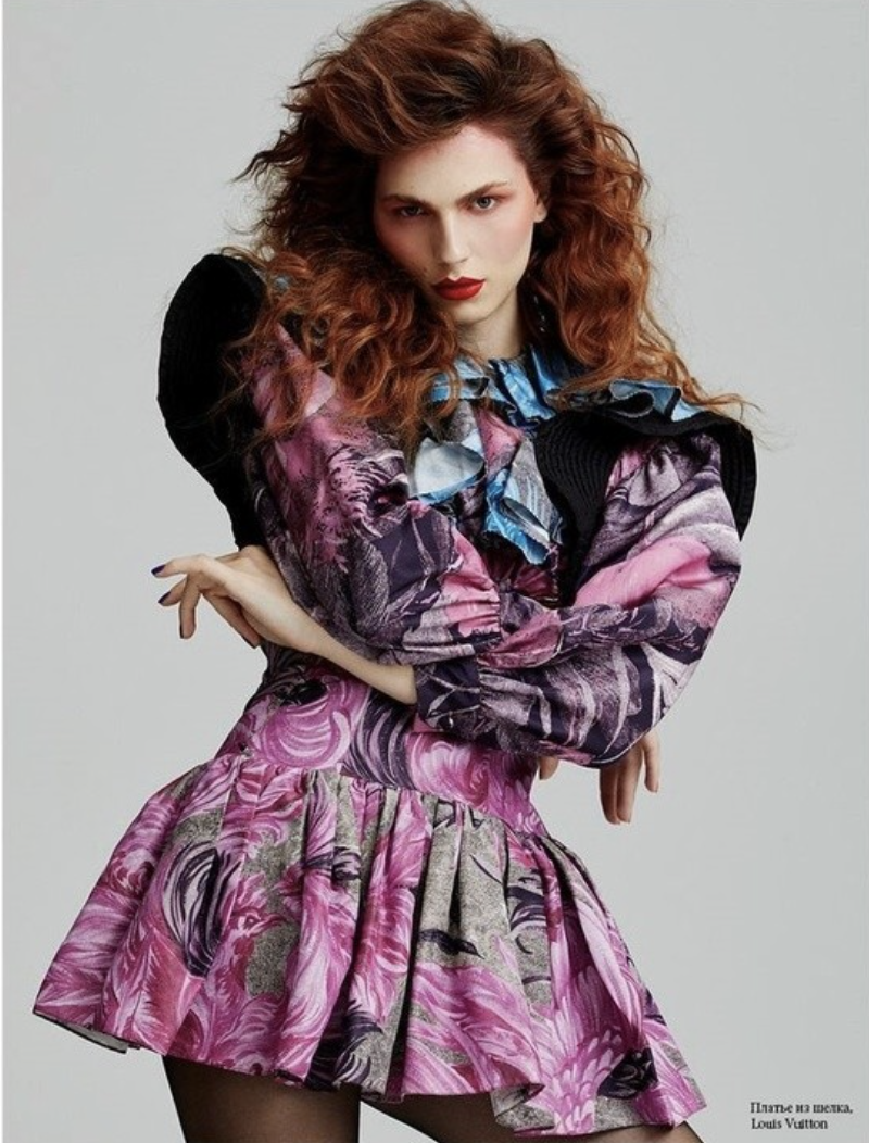 A woman with curly red hair poses in a colorful floral-print dress against a neutral background in a fashion editorial photograph.