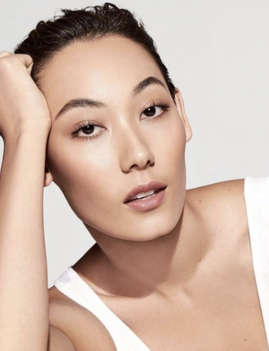 Close-up studio portrait of a young Asian woman with dark hair, resting her head on her hand, wearing a white top against a pale background.