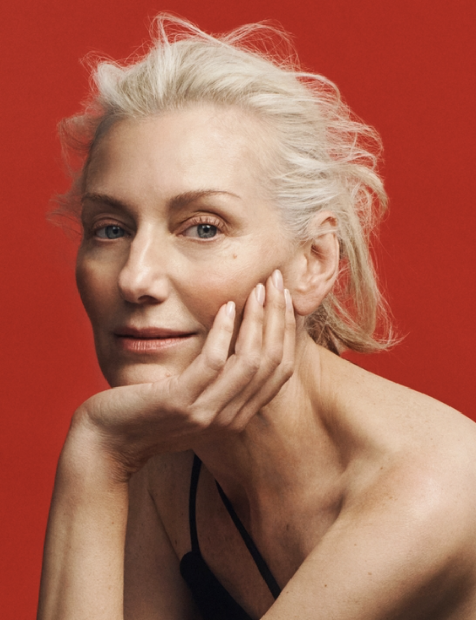 Close-up portrait of an adult woman with silver hair resting her chin on her hand against a vivid red background in a photo studio.