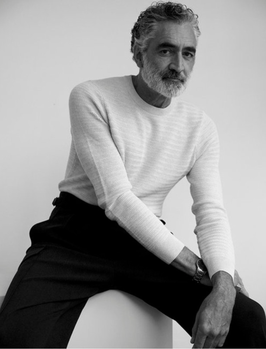 Monochrome portrait of a mature male model with grey hair and beard in a light sweater and dark trousers, sitting indoors against white wall.