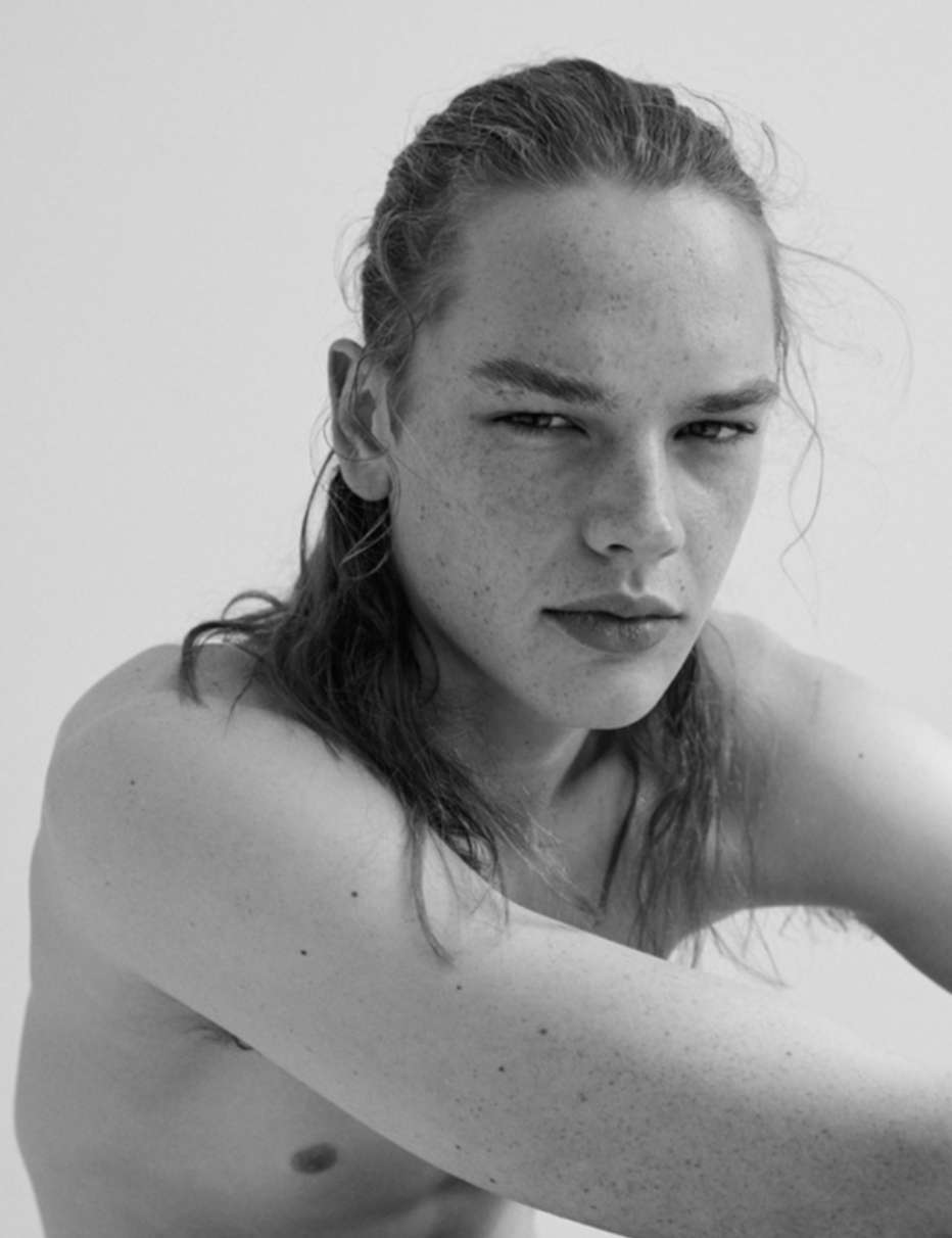 Black and white portrait of an adult male with long hair pulled back, fair skin, freckles, and bare shoulders against a white backdrop.