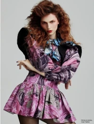 Model with voluminous red hair wearing a patterned pink and purple ruffled dress against a grey studio background