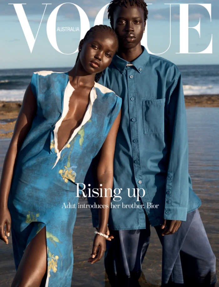 Magazine cover featuring two Black models standing in shallow water near a rocky coast under clear sky.