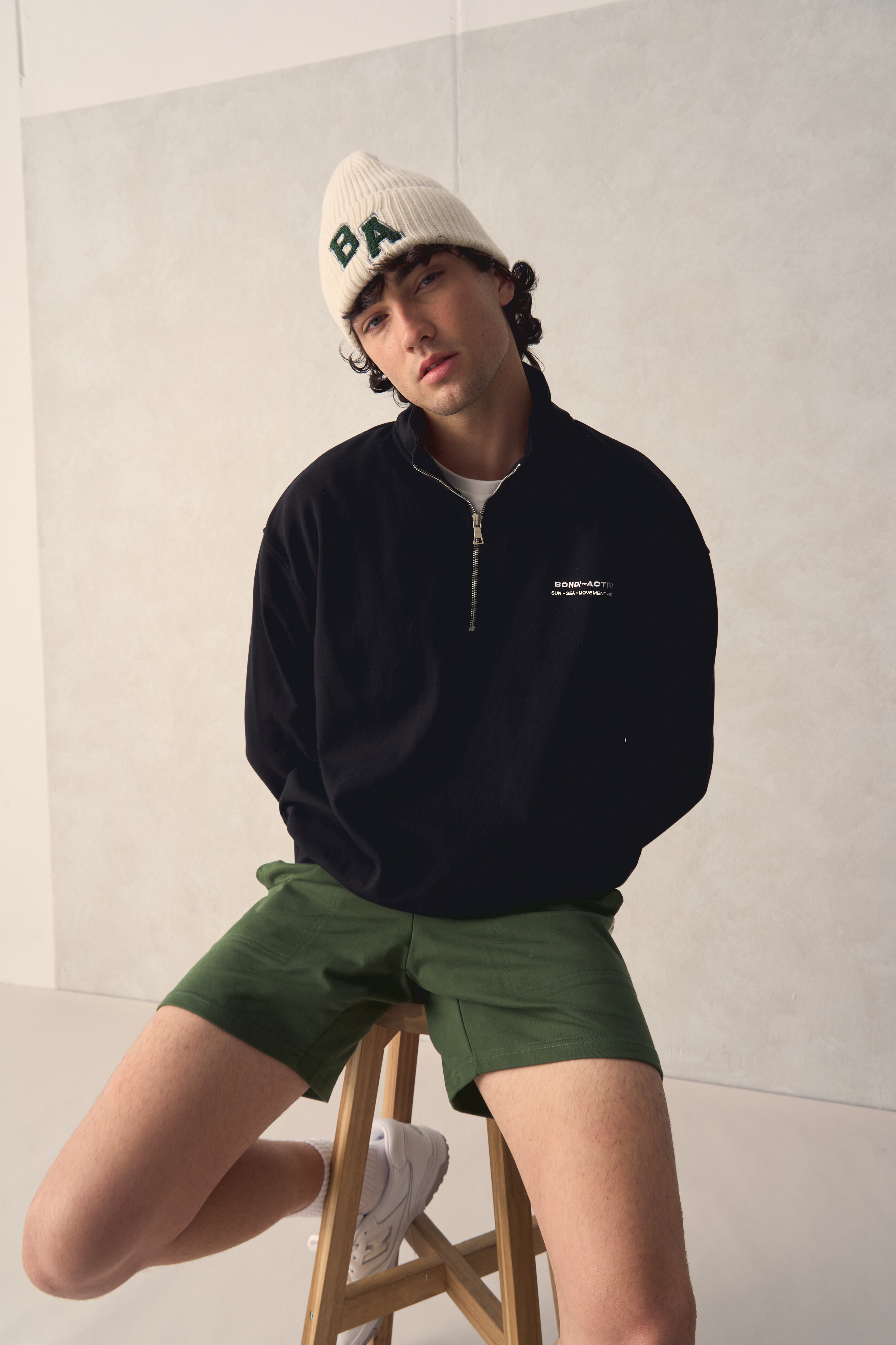 Young adult male poses indoors wearing a black sweater, green shorts, and a cream-colored beanie, sitting on a wooden stool. 