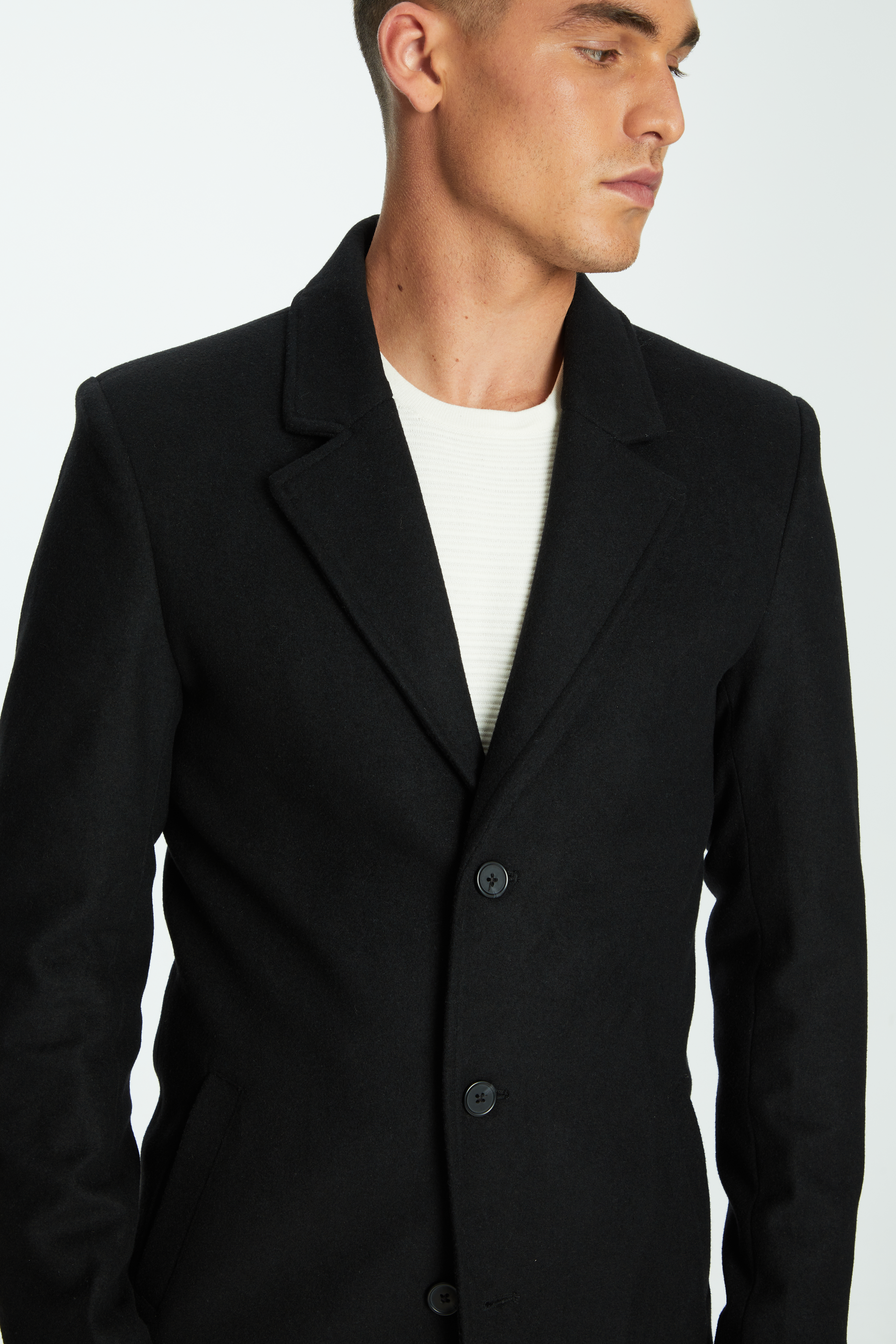 Adult male wearing a black overcoat and white shirt in a studio setting, showcasing fashion and style.