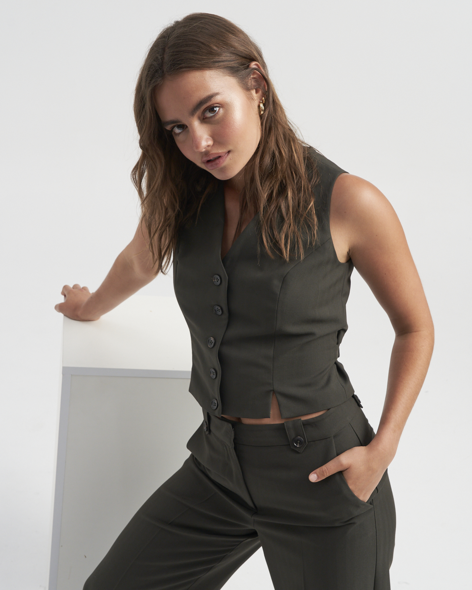 A woman with brown hair poses in a photo studio wearing a dark green buttoned vest and matching pants. She leans against a white cube.