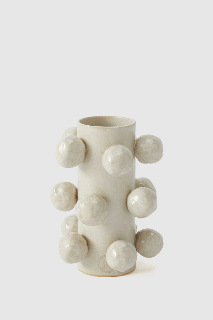 Cylindrical ceramic vase with attached spherical clay balls, studio shot against white background, featuring neutral tones and rough textures.