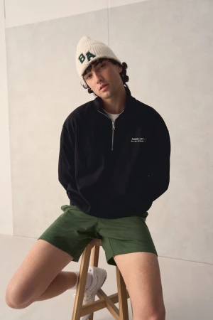 Young man wearing a cream beanie, black zip sweatshirt, and green shorts seated on a wooden stool in a studio setting.