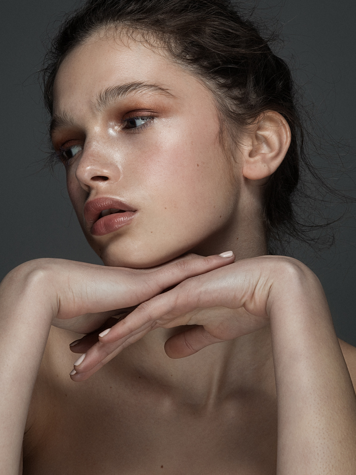 Close-up portrait of a young adult woman with hands near her face, soft makeup, and an expressive gaze in a studio setting.