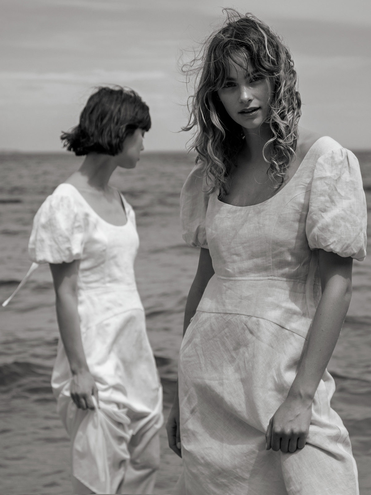 Two women in white dresses pose in the ocean under an overcast sky; one gazes at the camera, the other looks away in a black-and-white photo.