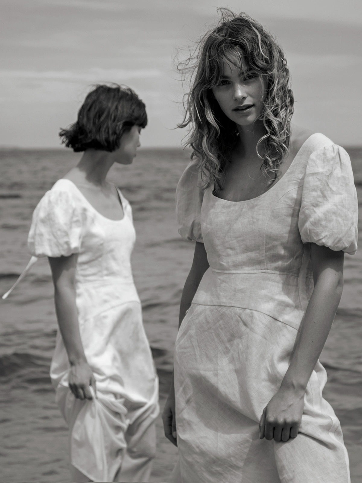 Two women in white dresses standing by the water in a black and white photograph by Peter Soulis.