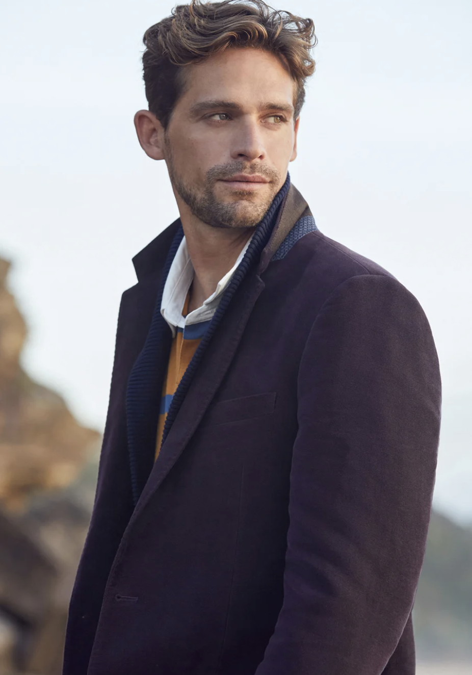 Portrait of a handsome man in a purple corduroy blazer and striped shirt, standing against a natural coastal background.